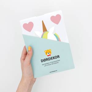 Dørdekor for barnerom med enhjørning og hjerter, holdt i en hånd med gul neglelakk. Dekoren er pakket i et lyseblått omslag med et bjørneikon. Ideell for å tilføre et lekent preg på døren til barnerommet.