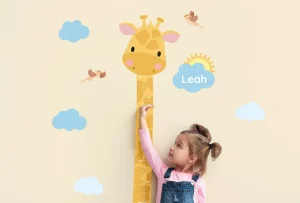 Barnet måler høyden sin ved en fargerik giraff-veggdekorasjon med innskriften Leah, omkranset av søte skyer og fugler. Denne veggdekorasjonen er perfekt for barnerommet, og bidrar til en inspirerende og lekende atmosfære.