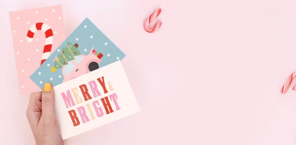 Hånd som holder fargerike bursdagskort med julemotiver og teksten “Merry Bright” og "Ho Ho Ho" på en julegave.