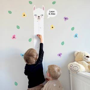 To barn måler høyden sin ved hjelp av en alpaka-formet høydeveggdekor i et barnerom, omgitt av fargerike blomster og blader. Veggdekoren har personlige navn og skaper en leken og funksjonell atmosfære.