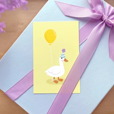 Alt-tekst: Illustrasjon av en glad and med en gul ballong, som står foran en gave pakket inn i blått papir med en lilla sløyfe. Dette bursdagskortet passer perfekt til barn og kan brukes til å feire spesielle anledninger.