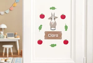 Fargerik dørdekor for barn med en koselig geit og navnet Clara på en treplate, omringet av røde epler og grønne blader, perfekt for å personliggjøre barnerommet.