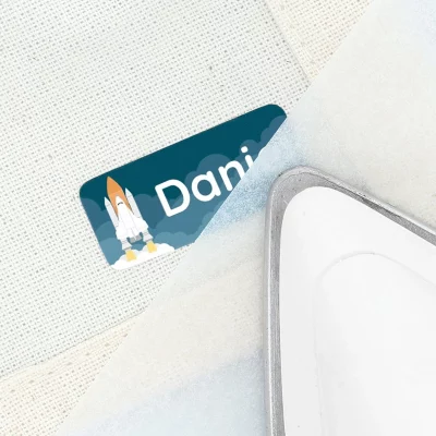 Navnelapp med navnet 'Daniel' og en rakettillustrasjon, delvis skjult bak et strykesett. Perfekt for å merke personlige eiendeler og gjøre dem unike.