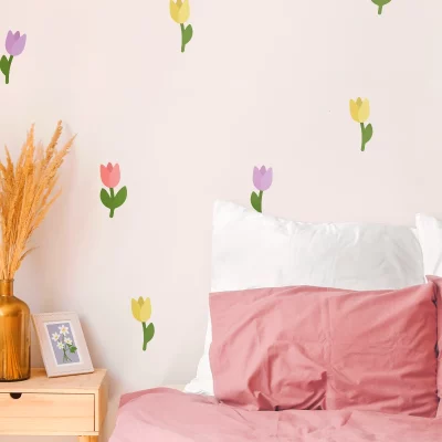 Interiørbilde av et barnerom med en vegg dekorert med fargerike blomstervegger. I forkant sees en blomsterpotte med tørkede blomster og en ramme med en illustrasjon. Bildet viser en koselig atmosfære og illustrerer hvordan veggdekor kan gi liv til et barnerom.