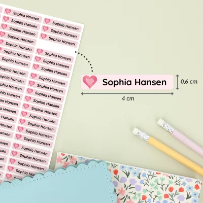 Navnelapper med hjerte og egen tekst, designet for personlig tilpasning. Hver etikett måler 4 cm x 0,6 cm og er perfekt for å merke barns eiendeler. I bakgrunnen er det fargeglade skrivesaker og dekorative notatbøker, ideelt for barnerommet.