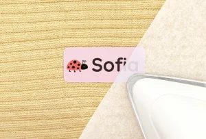 Bilde av en navnelapp i rosa med navn Sofia og en søt marihøneillustrasjon. Lappen ligger på en strukturert bakgrunn av beige stoff, ved siden av et strykejern. Dette produktet er perfekt for personlig tilpasning av barns eiendeler.