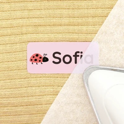 Bilde av en navnelapp i rosa med navn Sofia og en søt marihøneillustrasjon. Lappen ligger på en strukturert bakgrunn av beige stoff, ved siden av et strykejern. Dette produktet er perfekt for personlig tilpasning av barns eiendeler.