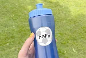 En person holder en blå drikkeflaske med en tilpasset navnelapp som har teksten Felix og en fotballdesign. Bakgrunnen viser frodig grønt gress, som gir en sommerlig og aktiv atmosfære. Denne flasken er perfekt til bruk under lek og sport, og navnelappen sikrer at flasken enkelt kan identifiseres.