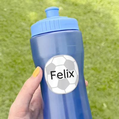 En person holder en blå drikkeflaske med en tilpasset navnelapp som har teksten Felix og en fotballdesign. Bakgrunnen viser frodig grønt gress, som gir en sommerlig og aktiv atmosfære. Denne flasken er perfekt til bruk under lek og sport, og navnelappen sikrer at flasken enkelt kan identifiseres.