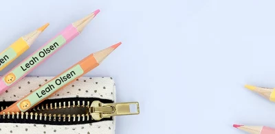 Fargerike fargeblyanter merket med navnelapper Leah Olsen ligger sammen med en linjal og en liten penal. Dette bildet illustrerer kreative produkter til barn, inkludert navnelapper for personliggjøring av skoleutstyr.