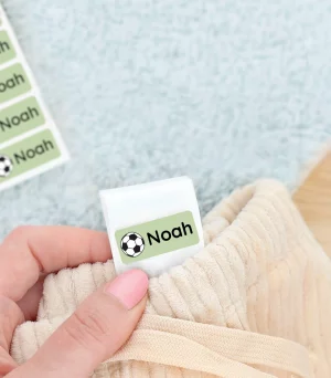 Bilde av en hånd som holder en navnelapp med navnet Noah og et fotball design, plassert pp en vaskelapp i en bukse. I bakgrunnen sees et par grønne barnesko og et mykt blått teppe. Navnelapper for barn, perfekte for å organisere klær og sko.