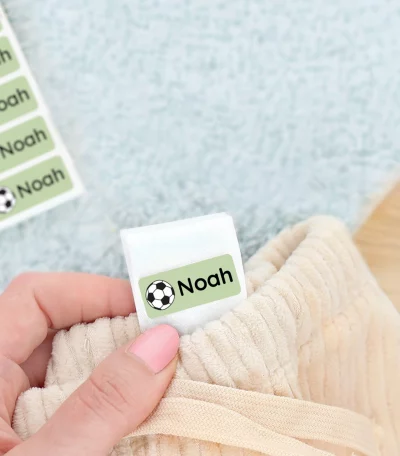 Bilde av en hånd som holder en navnelapp med navnet Noah og et fotball design, plassert pp en vaskelapp i en bukse. I bakgrunnen sees et par grønne barnesko og et mykt blått teppe. Navnelapper for barn, perfekte for å organisere klær og sko.