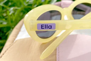 Gule solbriller med navnelapp som viser navnet Ella. Brillene ligger på en lys, teksturert overflate med grønt gress i bakgrunnen, som gir et sommerlig preg. Dette produktet er perfekt for å personliggjøre barnas tilbehør og gjøre dem mer gjenkjennelige.