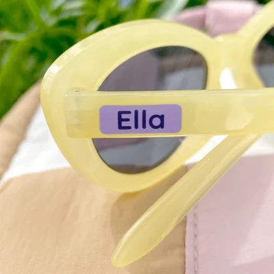Gule solbriller med navnelapp som viser navnet Ella. Brillene ligger på en lys, teksturert overflate med grønt gress i bakgrunnen, som gir et sommerlig preg. Dette produktet er perfekt for å personliggjøre barnas tilbehør og gjøre dem mer gjenkjennelige.
