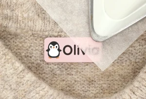 Bildet viser en navnelapp med design av en pingvin, som har navnet Olivia trykt på en rosa bakgrunn. Lappen er plassert på en myk, beige overflate, og en sylinderformet gjenstand i metall ligger over et lag med papir, klar til bruk. Dette produktet er perfekt for å merke klær eller andre eiendeler til barn.