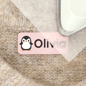 Bildet viser en navnelapp med design av en pingvin, som har navnet Olivia trykt på en rosa bakgrunn. Lappen er plassert på en myk, beige overflate, og en sylinderformet gjenstand i metall ligger over et lag med papir, klar til bruk. Dette produktet er perfekt for å merke klær eller andre eiendeler til barn.