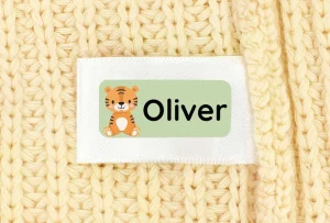 Bilde av en navnelapp med teksten Oliver og en søt tegning av en tiger. Lappen er festet til et mykt, gyllent strikket tekstil. Lappene våre er perfekte for personlig merking av klær og eiendeler til barn.