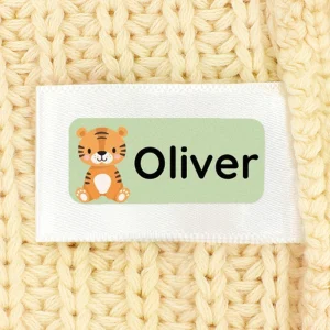 Bilde av en navnelapp med teksten Oliver og en søt tegning av en tiger. Lappen er festet til et mykt, gyllent strikket tekstil. Lappene våre er perfekte for personlig merking av klær og eiendeler til barn.
