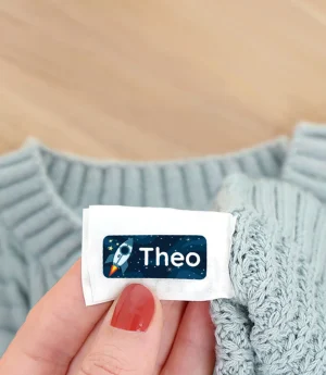 Bilde av en hånd som holder en navnelapp med navnet Theo på en lys, strikket genser. I bakgrunnen ses en beige støvel og fargerike høstblader på en lys treoverflate. Dette bildet illustrerer bruk av navnelapper, perfekt for å merke klær til barn.